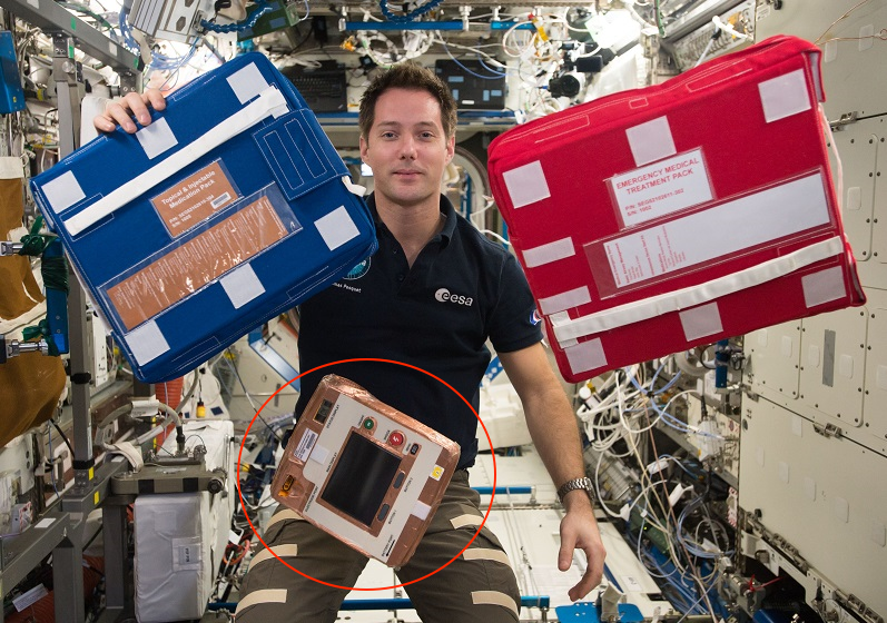 ESA astronaut Thomas Pesquet with the automated external defibrillator floating below two medical packspng